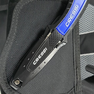 Puede incluir: Un cuchillo de buceo negro con mango azul y la palabra "CRESSI" impresa en él. El cuchillo está sujeto a un chaleco de buceo negro.