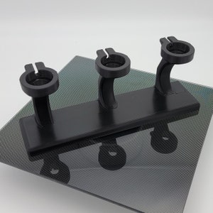 May include: A black plastic stand with three holders for displaying items. The stand has a rectangular base and three vertical arms, each with a circular opening at the top.