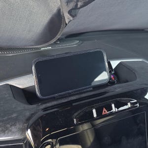 May include: A black smartphone in a clear case is mounted on a car dashboard. The phone is positioned horizontally, displaying a dark screen. The dashboard is black and gray, with a few buttons and a screen visible below the phone.