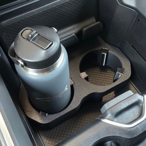 May include: A gray stainless steel water bottle with a black lid and a black fish logo is in a black cup holder in a car. The cup holder is in the center console of the car.
