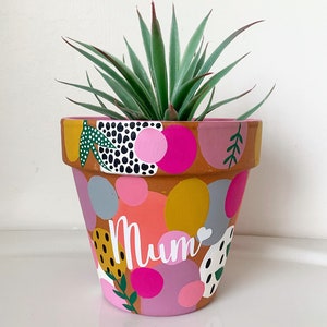 May include: A terracotta planter painted with pink, yellow, and green abstract shapes and the word "Mum". A green succulent plant is growing in the planter.