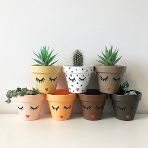 May include: Seven terracotta flower pots with painted faces. Each pot has a different colour and features black eyelashes and a red heart. The pots are arranged in a row on a white surface.