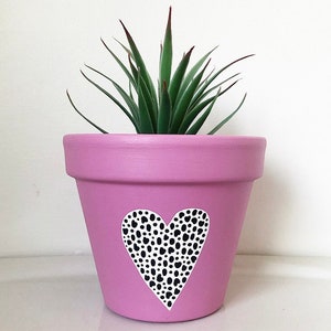 May include: A pink terracotta planter with a white heart design featuring black polka dots. The planter is filled with a green succulent plant.