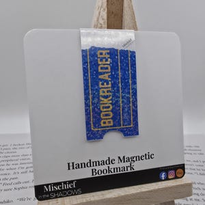 May include: A handmade magnetic bookmark with the word "BOOKREADER" in gold lettering on a blue background. The bookmark is attached to a white card, and the words "Handmade Magnetic Bookmark" are printed below. The bookmark is displayed on a small wooden easel.
