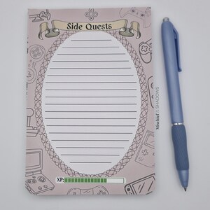 May include: A light pink notepad with the text "Side Quests" and a decorative oval border. The notepad has lined pages and is decorated with video game controller illustrations. A blue pen rests beside the notepad.