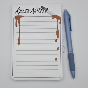May include: A white notepad with the text "Killer Notes" and a dripping blood design, accompanied by a blue pen. The notepad has lined pages and a black border. The pen is light blue with a dark blue grip.