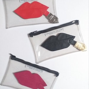 Può includere: Tre pochette trasparenti in plastica con cerniera, con un design di labbra rosse, nere e rosa e rossetto. Ogni pochette ha un'etichetta che recita "Handbags by Ann-Marie Made in London."