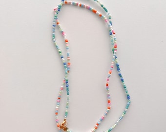 May include: A necklace made of small, multi-coloured glass seed beads in shades of pink, orange, blue, green, and clear. The strand features a gold-toned metal lobster claw clasp at the ends, allowing the beads to form a continuous loop.