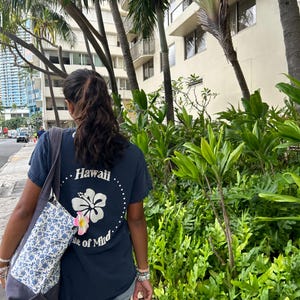 Op de afbeelding: Een donkerblauw T-shirt met korte mouwen voorzien van een witte hibiscusbloem-afbeelding, een gestippelde cirkel en de tekst Hawaii State of Mind op de achterkant, gecombineerd met een grijs-witte bloemrijke tas met een roze-witte plumeria.