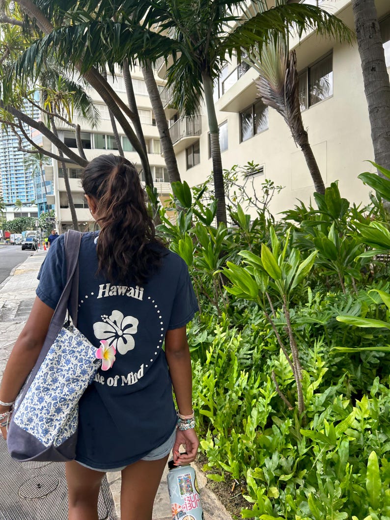 Op de afbeelding: Een donkerblauw T-shirt met korte mouwen voorzien van een witte hibiscusbloem-afbeelding, een gestippelde cirkel en de tekst Hawaii State of Mind op de achterkant, gecombineerd met een grijs-witte bloemrijke tas met een roze-witte plumeria.