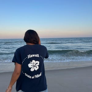 Op de afbeelding: Een marineblauw t-shirt met korte mouwen en een witte afbeelding op de achterkant. Het ontwerp bevat een centrale hibiscusbloem omringd door een stippellijn, met de tekst "Hawaii" bovenaan en "State of Mind" onderaan in witte letters.