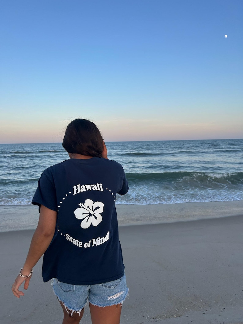 Op de afbeelding: Een marineblauw t-shirt met korte mouwen en een witte afbeelding op de achterkant. Het ontwerp bevat een centrale hibiscusbloem omringd door een stippellijn, met de tekst "Hawaii" bovenaan en "State of Mind" onderaan in witte letters.