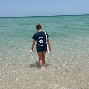 Op de afbeelding: Een donkerblauw t-shirt met korte mouwen en een witte afbeelding op de achterkant. De afbeelding toont een hibiscusbloem in een gestippelde cirkel, met de tekst Hawaii State of Mind in een cirkel rondom de bloem gedrukt.