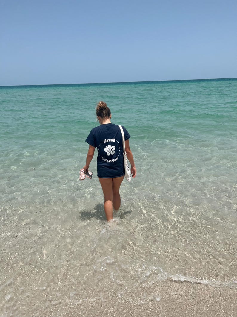 Op de afbeelding: Een donkerblauw t-shirt met korte mouwen en een witte afbeelding op de achterkant. De afbeelding toont een hibiscusbloem in een gestippelde cirkel, met de tekst Hawaii State of Mind in een cirkel rondom de bloem gedrukt.