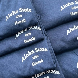 Op de afbeelding: Een stapel opgevouwen marineblauwe T-shirts, elk voorzien van witte tekst met de opdruk Aloha State, 1959 en Hawaii. De shirts zijn in een stapel gerangschikt, waardoor het consistente grafische ontwerp op het textieloppervlak zichtbaar is.