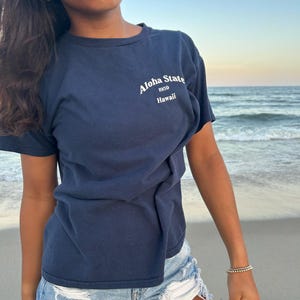 Op de afbeelding: Een marineblauw t-shirt met korte mouwen en witte tekst op de borst met de tekst Aloha State 1959 Hawaii. Het shirt heeft een ronde hals en wordt gecombineerd met een lichtblauwe spijkerbroek met een gerafelde zoom en witte zakvoering.
