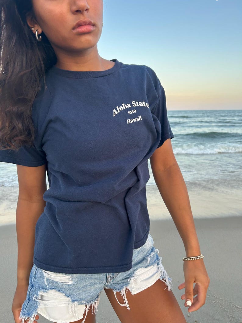 Op de afbeelding: Een marineblauw t-shirt met korte mouwen en witte tekst op de borst met de tekst Aloha State 1959 Hawaii. Het shirt heeft een ronde hals en wordt gecombineerd met een lichtblauwe spijkerbroek met een gerafelde zoom en witte zakvoering.