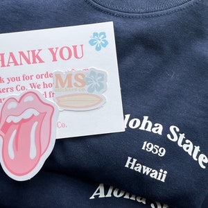 Op de afbeelding: Een donker marineblauw t-shirt met witte tekst Aloha State 1959 Hawaii. Inbegrepen is een witte bedankkaart met roze tekst, een lichtblauwe hibiscusbloem en twee stickers: een met een roze tongafbeelding en een met de tekst MS stickers co.