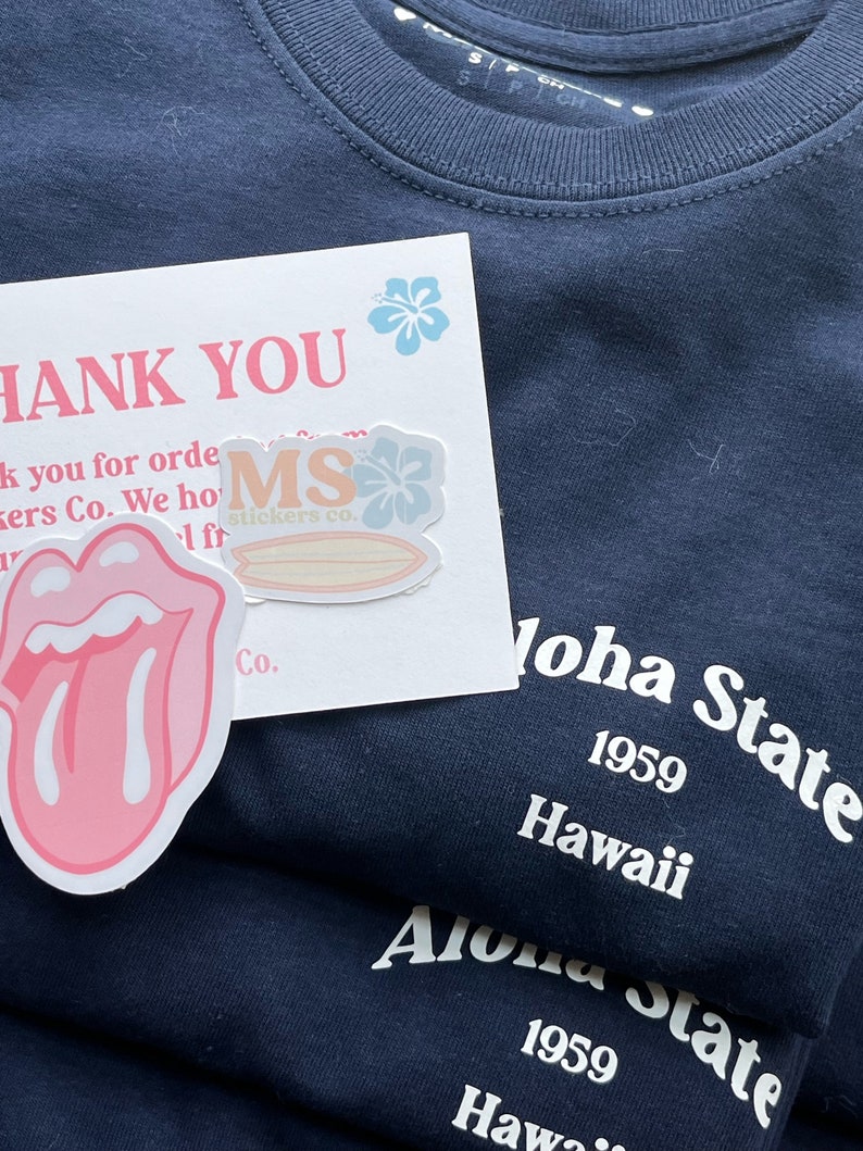 Op de afbeelding: Een donker marineblauw t-shirt met witte tekst Aloha State 1959 Hawaii. Inbegrepen is een witte bedankkaart met roze tekst, een lichtblauwe hibiscusbloem en twee stickers: een met een roze tongafbeelding en een met de tekst MS stickers co.