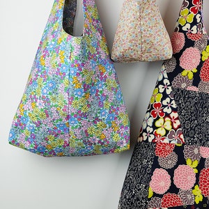 May include: Three fabric tote bags with floral patterns hang against a white wall. The bags feature various floral designs in blue, pink, yellow, and red. The bags are different sizes and shapes, with one having a patchwork design.