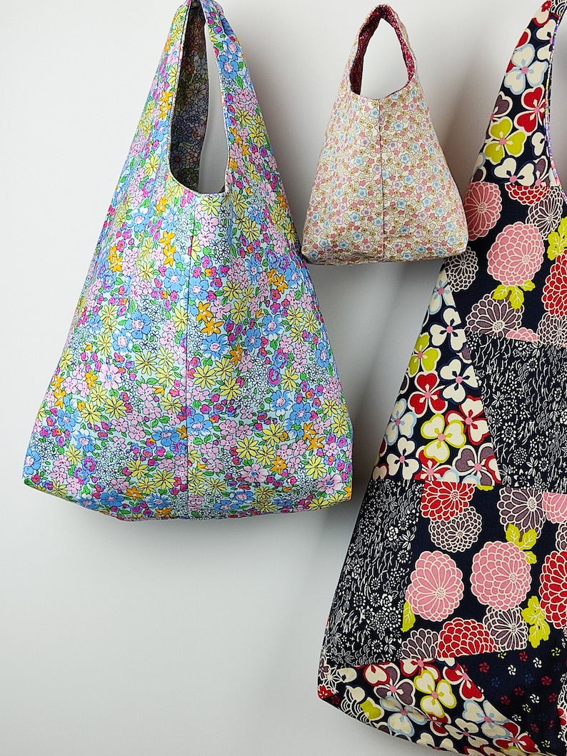 May include: Three fabric tote bags with floral patterns hang against a white wall. The bags feature various floral designs in blue, pink, yellow, and red. The bags are different sizes and shapes, with one having a patchwork design.