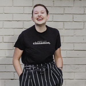 May include: A person wearing a black t-shirt with the text "Sweet Like Cinnamon" in white. They are smiling and wearing black and white striped pants.