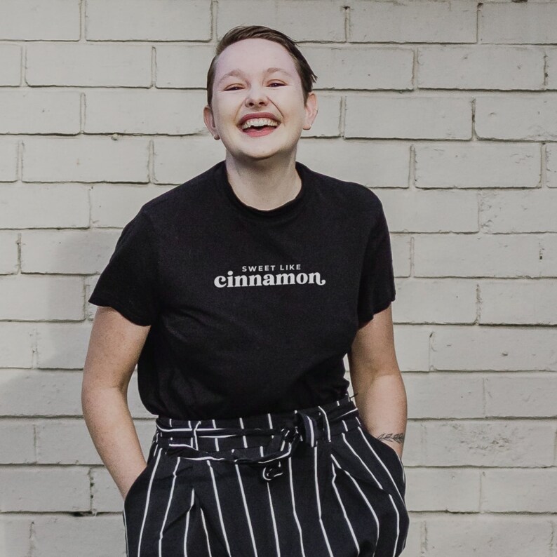May include: A person wearing a black t-shirt with the text "Sweet Like Cinnamon" in white. They are smiling and wearing black and white striped pants.