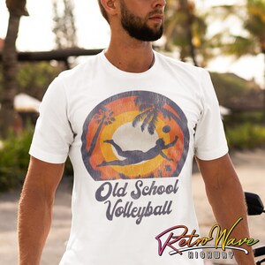 Op de afbeelding: Een wit T-shirt met een vintage-stijl grafische afbeelding van een persoon die volleybal speelt op een strand met palmbomen. De afbeelding is in tinten oranje, geel en rood. De tekst "Old School Volleyball" is onder de afbeelding gedrukt.