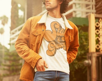 May include: A man wearing a brown corduroy jacket over a white t-shirt with the text "Stay Groovy" in a retro font. He is wearing blue jeans and has curly brown hair. He is wearing yellow-tinted sunglasses.