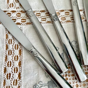 May include: Close-up of several silver butter knives. The knives have a simple, elegant design with a floral pattern at the base of the blade. The blades are shiny and reflect the light. The knives are arranged on a white lace tablecloth.