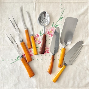 May include: A collection of vintage serving utensils, including forks, knives, a slotted spoon, and cake servers. The utensils feature stainless steel blades and orange and yellow handles. The items are arranged on a floral embroidered tablecloth.