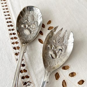 May include: Silver serving spoon and fork set. The spoon and fork heads feature an embossed design of fruit and leaves. The handles are long and slender. The set is displayed on a white, embroidered tablecloth.