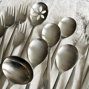May include: A collection of stainless steel silverware, including forks, spoons, and a slotted spoon with a floral design. The utensils are arranged on a white, textured surface, creating a classic and elegant aesthetic.