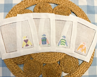 May include: Four white linen napkins with embroidered jockey silks in different colors. The silks are yellow and purple, pink and blue, green and yellow, and orange and pink.