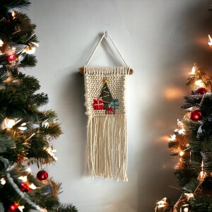 Puede incluir: Un tapiz de macramé blanco con un diseño de árbol de Navidad y regalos. El tapiz está hecho de fibras naturales y tiene una franja en la parte inferior.