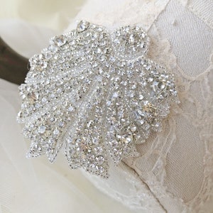 May include: A silver rhinestone applique in the shape of a leaf. The applique is made of small, clear rhinestones that are sewn onto a fabric base.