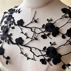 Könnte beinhalten: Eine schwarze Spitzenapplikation mit einem komplizierten Stickdesign aus blühenden Zweigen mit Blüten und Knospen. Das zarte florale Motiv ist in einem weitläufigen, organischen Muster angeordnet und zeigt detaillierte Nähte an jedem Blütenblatt und Stiel.
