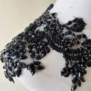 Può includere: Applicazione di perline nere con un design floreale. L'applicazione presenta varie forme di perline e paillettes nere, cucite su un tessuto a rete nero trasparente. Il design è esposto su un manichino.