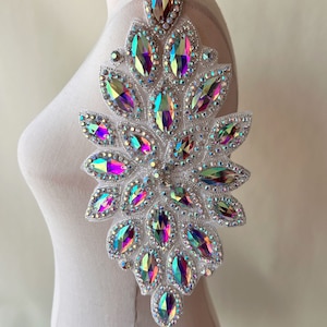 May include: A decorative applique with iridescent rhinestones in a floral pattern. The design features large, teardrop-shaped rhinestones in shades of pink, green, and blue. The rhinestones are set on a white fabric backing and are surrounded by smaller, clear rhinestones.