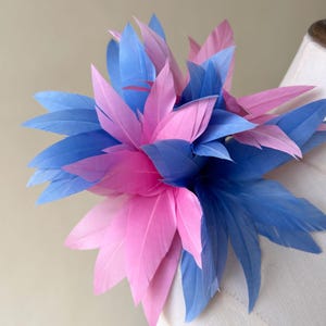 May include: A large, decorative flower made of layered feathers. The feathers are a mix of light blue and pink, arranged in a radial pattern. The flower is likely a millinery piece or a decorative accent for clothing or accessories.