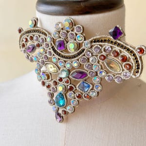 Pode incluir: Um colar choker dourado com design floral adornado com joias multicoloridas. O colar é adornado com pedras de cristal de vários tamanhos e formas, incluindo roxo, azul, verde e vermelho.