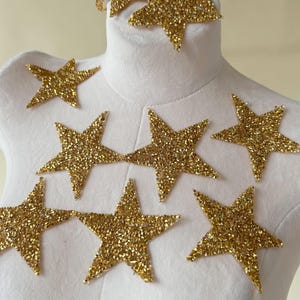 Puede incluir: Adornos de estrellas doradas con purpurina de varios tamaños dispuestos sobre una superficie blanca. Las estrellas son de cinco puntas y están cubiertas de purpurina dorada brillante. Estas estrellas decorativas son ideales para manualidades y añaden un toque de glamour a los proyectos.