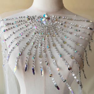 May include: A silver iridescent beaded and sequined shoulder piece with a large crystal centerpiece. The piece is designed to be worn as a decorative accessory.