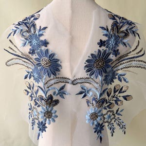 May include: An embroidered floral applique in shades of blue and silver. The design features detailed flowers, leaves, and sequins on a sheer, white fabric. Suitable for embellishing garments or craft projects. The measurements are in centimetres.