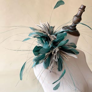 Puede incluir: Un fascinador de plumas verde azulado y blanco, exhibido en una cabeza de maniqu&iacute;. El accesorio presenta plumas verde azulado en capas y acentos de plumas blancas y grises, con acentos de plumas largas y finas verde azulado que se extienden hacia afuera. Adecuado para ocasiones especiales.