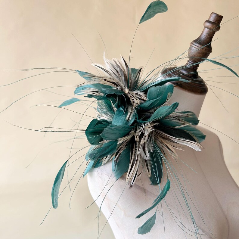 Puede incluir: Un fascinador de plumas verde azulado y blanco, exhibido en una cabeza de maniqu&iacute;. El accesorio presenta plumas verde azulado en capas y acentos de plumas blancas y grises, con acentos de plumas largas y finas verde azulado que se extienden hacia afuera. Adecuado para ocasiones especiales.
