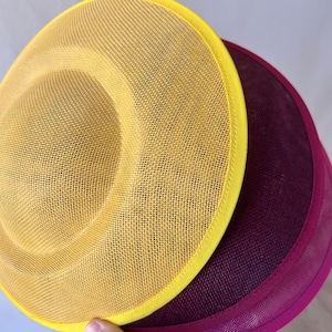 May include: Close-up of two hats. The top hat is yellow with a yellow trim. The bottom hat is a deep purple with a magenta trim. Both hats have a mesh-like texture.