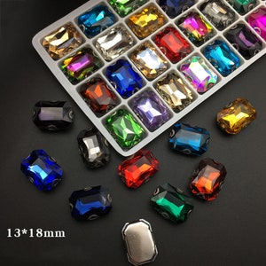 Puede incluir: Surtido de brillantes rectangulares de cristal de colores en una bandeja de plástico blanco. Los brillantes están dispuestos en filas y columnas, con cada brillante midiendo 13 mm por 18 mm. También hay brillantes sueltos sobre una superficie negra.