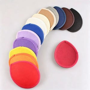 Puede incluir: Una colección de parches de tela en forma de lágrima en un espectro de colores que incluye rojo, naranja, amarillo, morado, lavanda, azul, crema, beige, gris, azul marino, marrón y negro, dispuestos en abanico con un parche rojo separado.