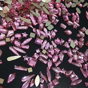 May include: Assortment of pink crystal rhinestones in various shapes and sizes, including stars, ovals, rectangles, and teardrops. The rhinestones are scattered on a black background.